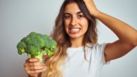 A young girl on white background is holding broccoli in her hand and is wondering what to do with it